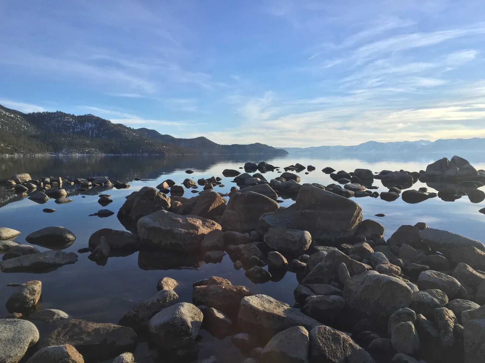 Rounded shoreline boulders reflected in Lake Tahoe, with snow-dusted wooded slopes across the water.